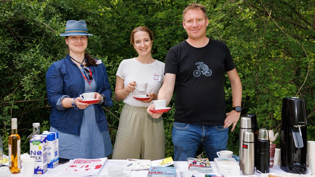 Drei Mitarbeiter:innen des Magistrats stehen hinter einem Tisch mit Infomaterial im Grünen, sie halten Kaffeetassen in der Hand. (Foto: Peter Rauchecker)
